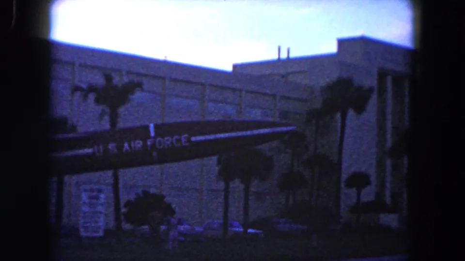 1960 Miami frame showing a U.S. Air Force aircraft display in front of a modern building, with palm trees, signs, and cars nearby.