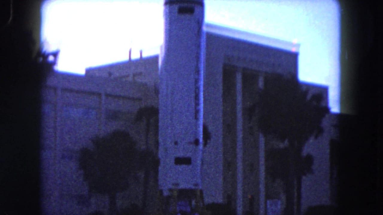 Rocket replica in front of a large building with palm trees and dark film borders.