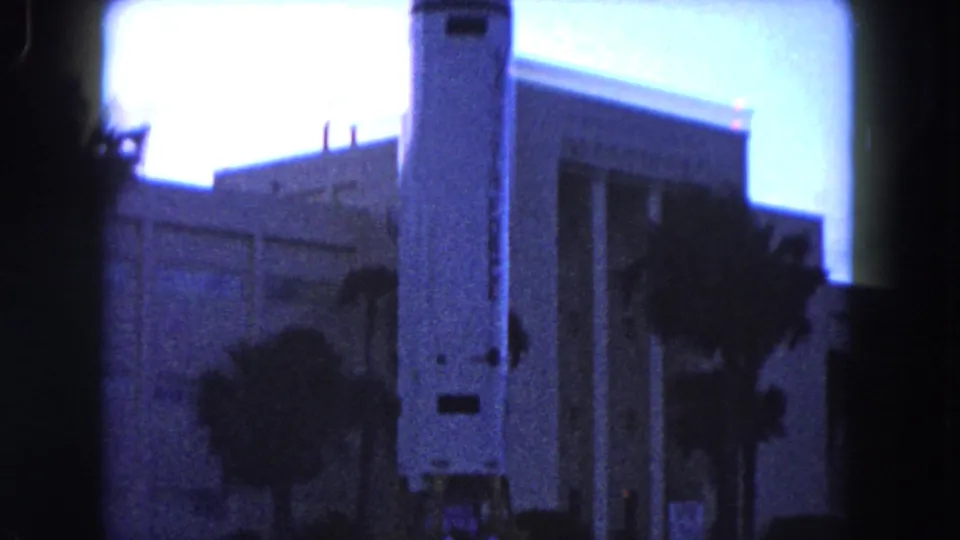 1960 Miami home-movie frame showing a rocket replica beside a large civic-looking building, with palm trees and blue-shifted film color.