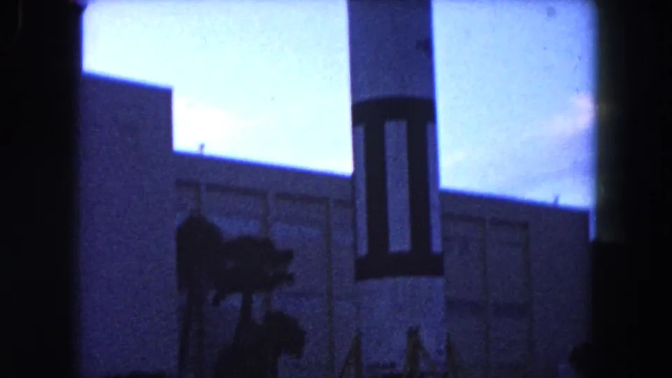 Wider 1960 Miami home-movie view of the rocket replica base in front of a large building, framed by palm trees and dark film edges.