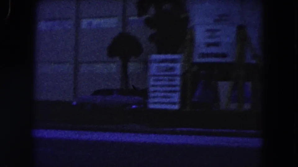 Low-light 1960 Miami street frame with temporary signs, a shadowed person, and a partial parked vehicle.
