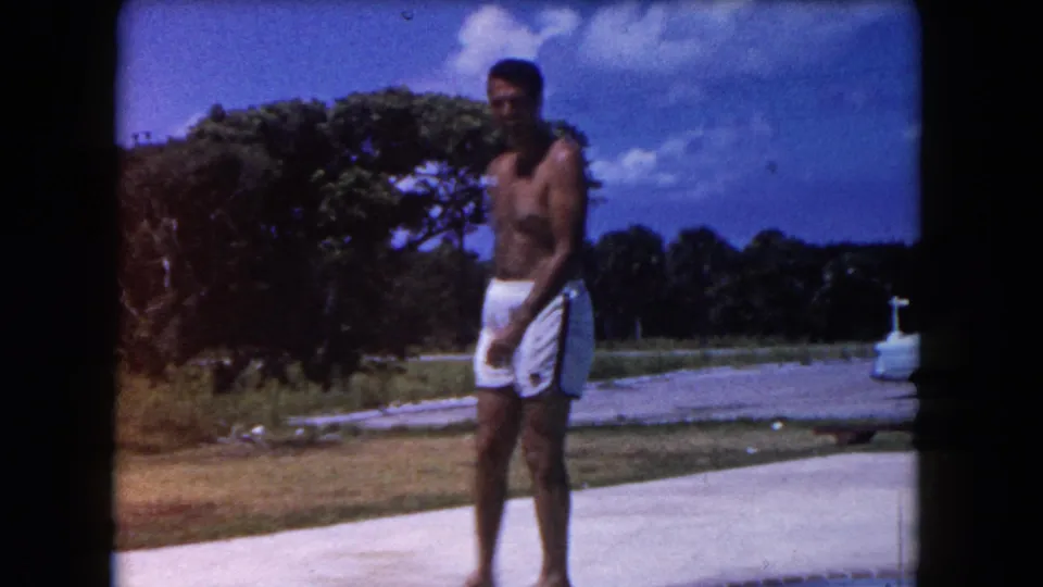 In 1960 Miami home-movie footage, a shirtless adult man in white trunks stands by a poolside patio under a blue sky.
