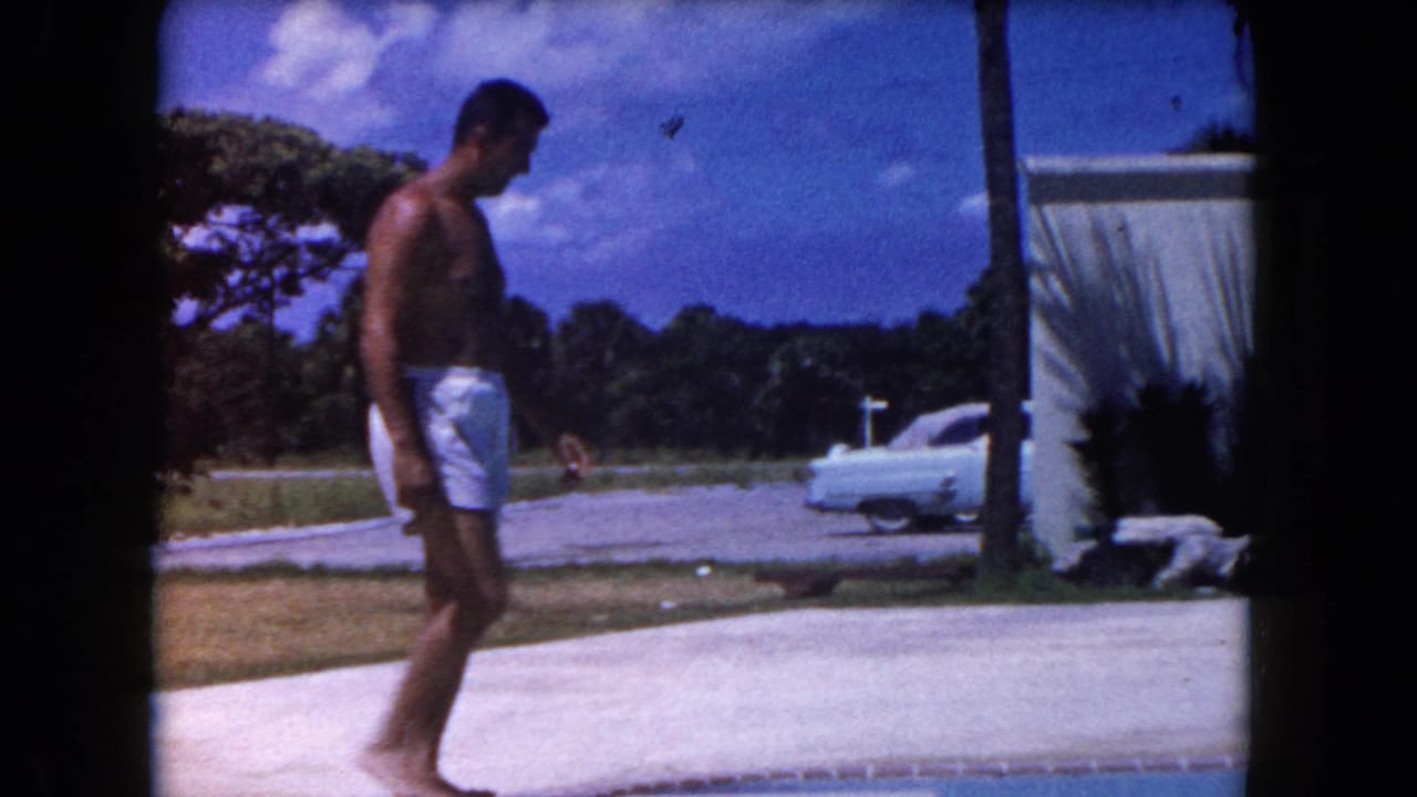 Shirtless man walks past a poolside patio with a white building and vintage car nearby.