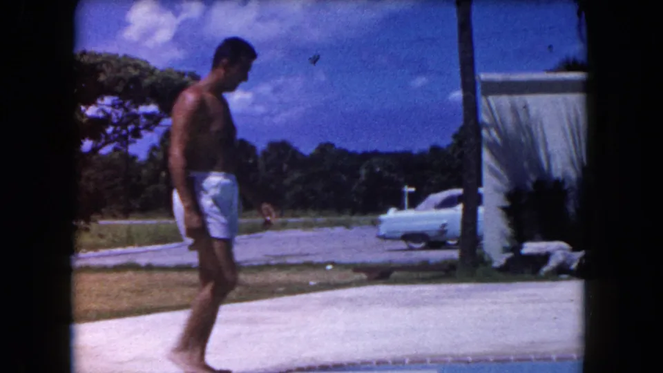 The man walks rightward along a Miami poolside patio, with a white building, palm, and light-colored vintage car visible.