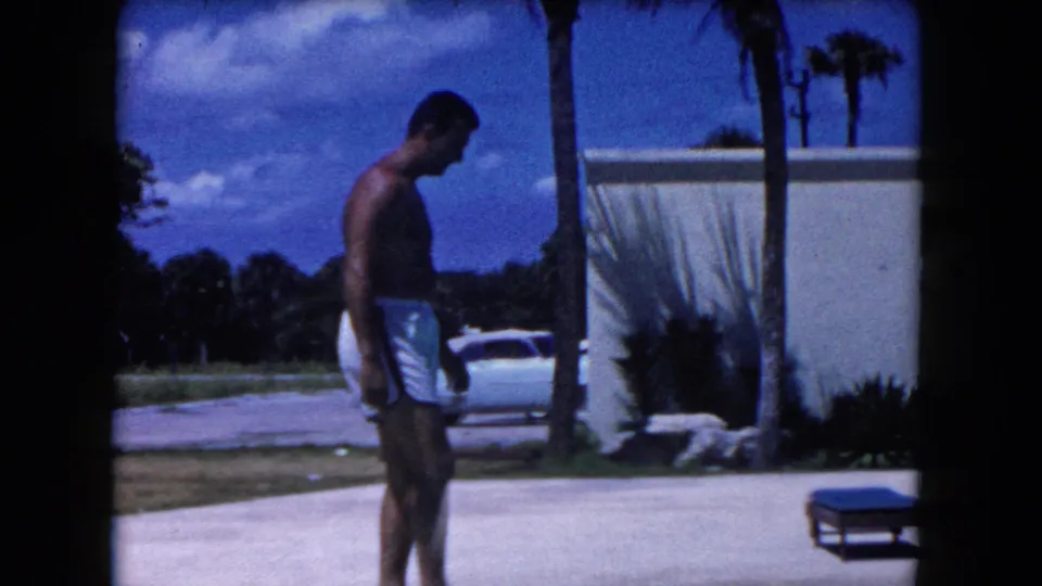 A shirtless man in white trunks moves across the poolside area, with a vintage car, palm trees, and modern white building behind him.