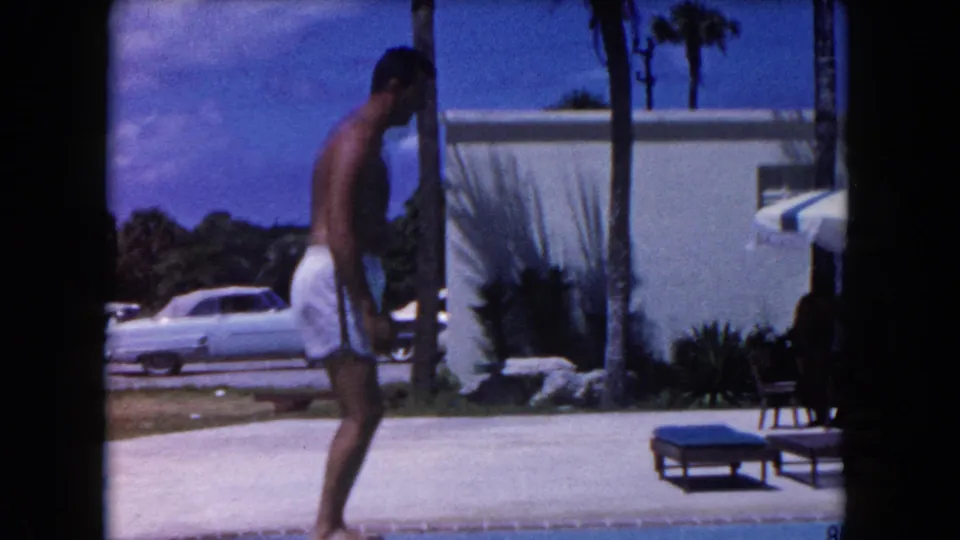 1960 Miami home-movie frame showing a man in white swim trunks walking poolside with a pale car, palm trees, and low building behind him.