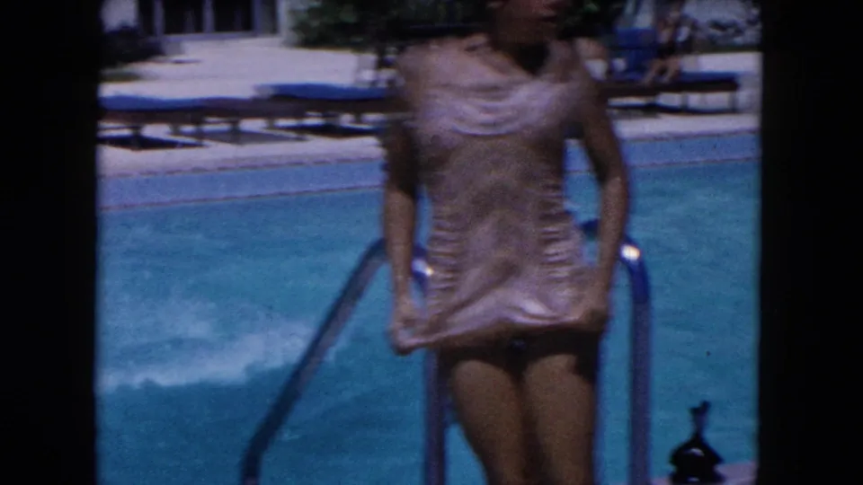 1960 Miami home-movie frame showing a woman in a vintage one-piece swimsuit entering the pool by a metal ladder beside blue lounge chairs.