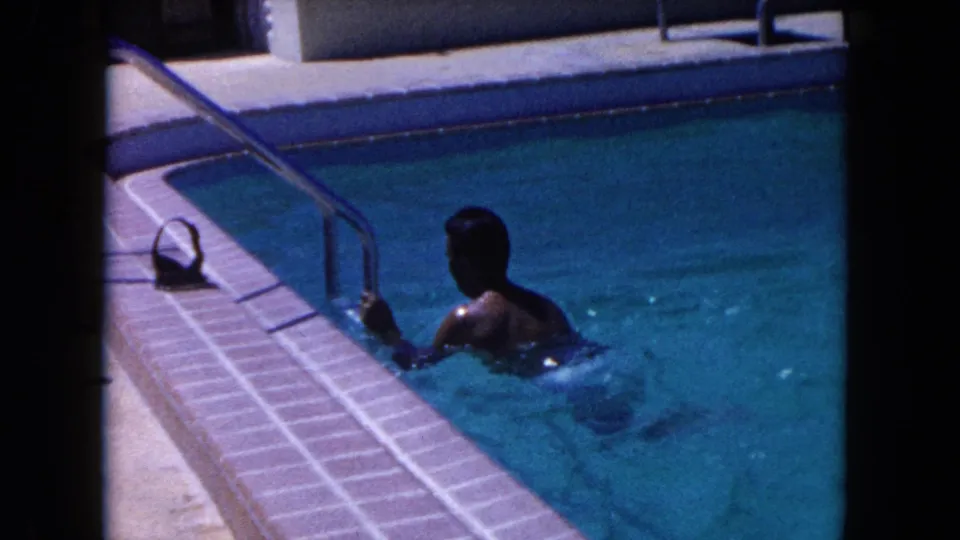 In 1960 Miami home-movie footage, a man in white swim shorts holds a pool ladder beside turquoise water and a tiled deck.