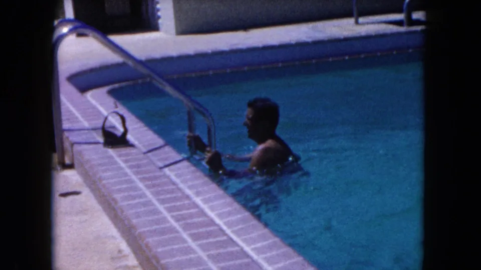The man remains near the ladder in the backyard pool, with bright water, tile coping, and deck visible in the 1960 Miami footage.