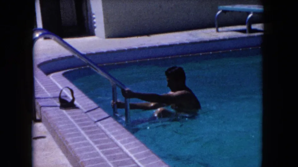 A man in white swim shorts reaches for the metal ladder in a sunlit backyard pool, seen in grainy 8mm Miami footage from 1960.