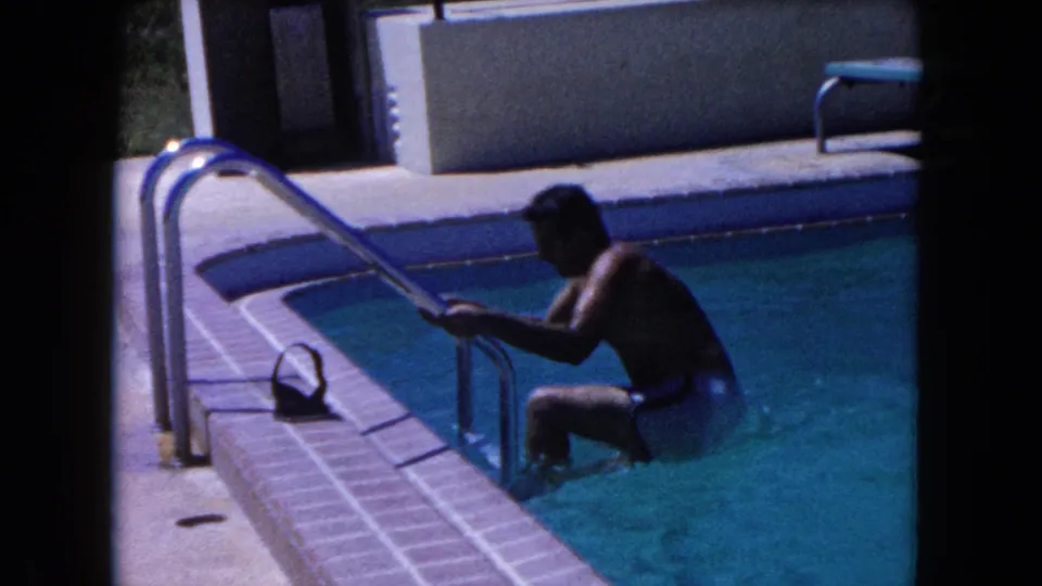 The man climbs the metal ladder from the backyard pool, with white swim shorts, tiled coping, and deck visible in the 1960 Miami home movie.