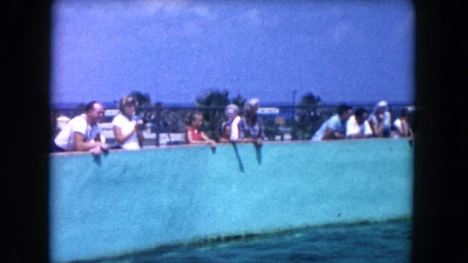 1960 Miami home-movie frame showing adults and children watching over a turquoise marine pool wall.