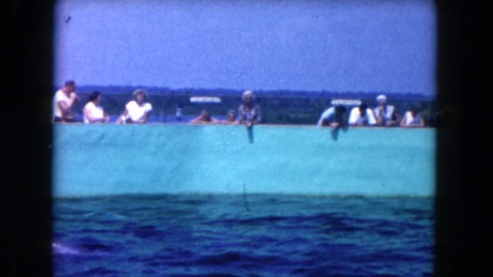 1960 Miami-area 8mm frame showing people in light clothing behind a turquoise wall above rippling water.