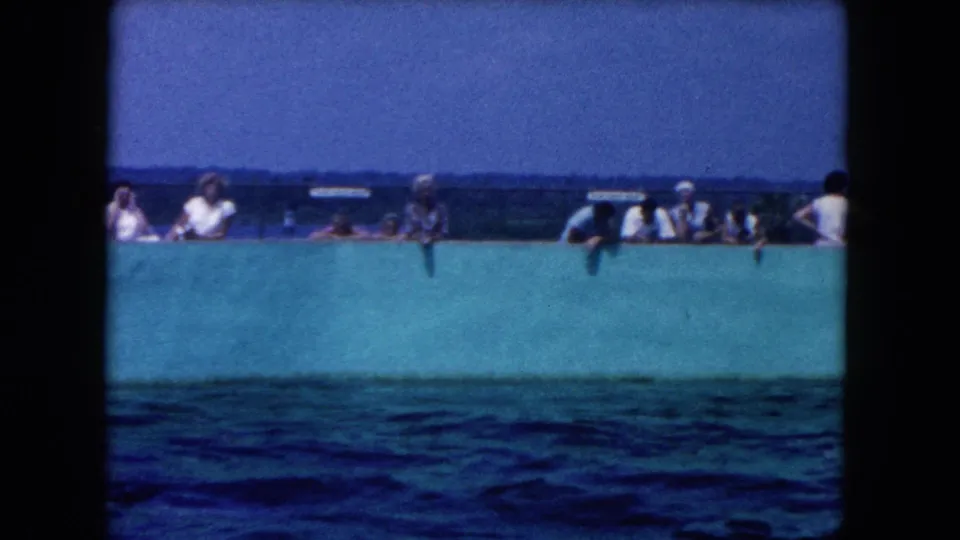 Miami-area home-movie frame with spectators along a turquoise wall and rippling water in the foreground.