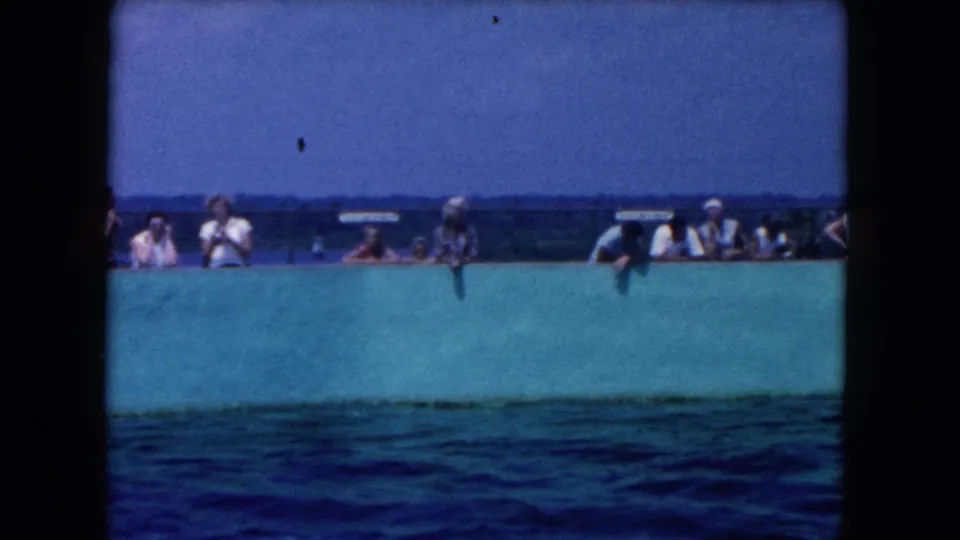 Soft 8mm frame of people near a turquoise dam or pool wall, with broad blue water in front.