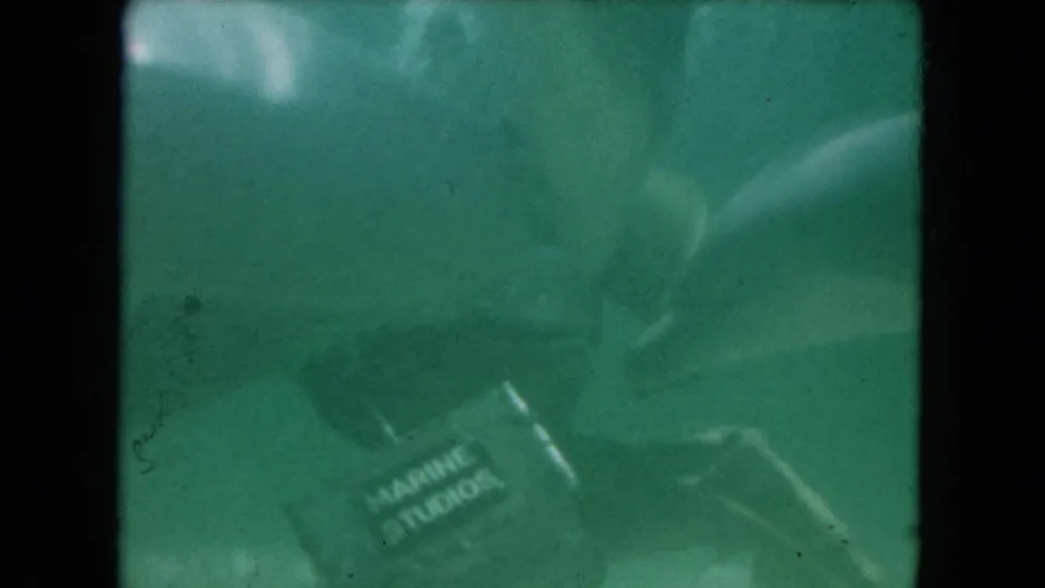 1960 Miami underwater 8mm frame showing a scuba diver near a Marine Studios-labeled container in murky green water.