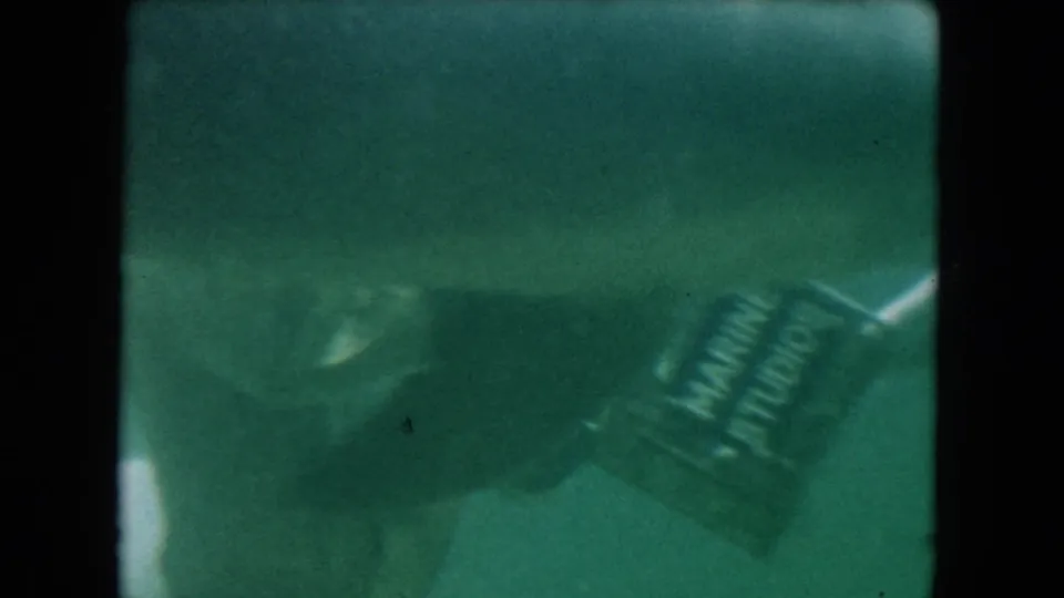 1960 Miami underwater 8mm frame with a helmeted diver holding a box near dolphins in soft green water.