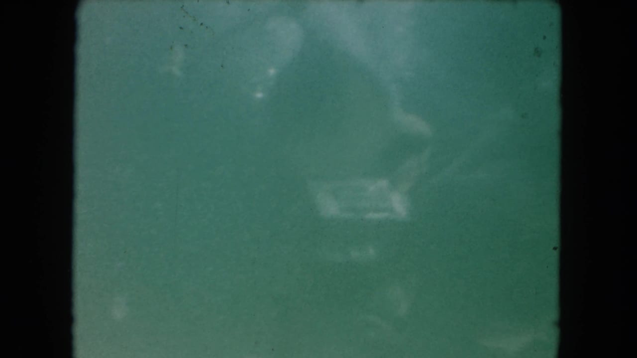 Hazy 1960 Miami pool footage shows indistinct fish shapes suspended in green-blue water on 8mm film.