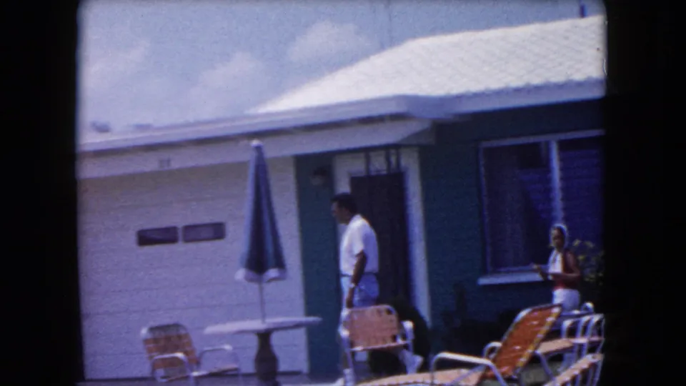 In a 1960 Miami home-movie frame, a man in light clothes walks near patio chairs and a closed blue umbrella outside a modest house.