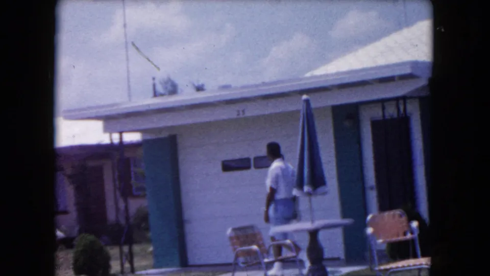 In Miami, 1960, a man in a white shirt and light shorts walks past patio furniture outside a low house with green trim.