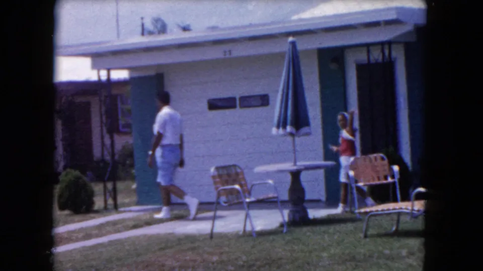 A 1960 Miami home-movie frame shows a man crossing the yard as a child in red and white stands near the doorway of the house.
