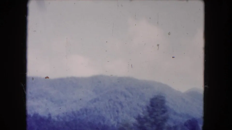 1960s 8mm home-movie frame showing distant wooded mountain ridges under an overcast sky, with aged blue-purple color shift.