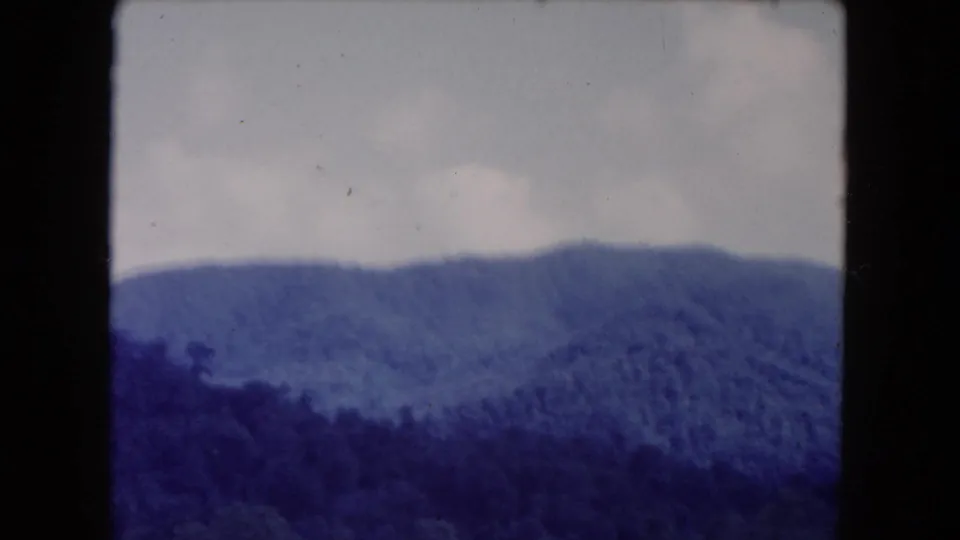 A wooded mountain ridge fills the lower frame while the pale sky and aged 8mm texture show dust, scratches, and color drift.