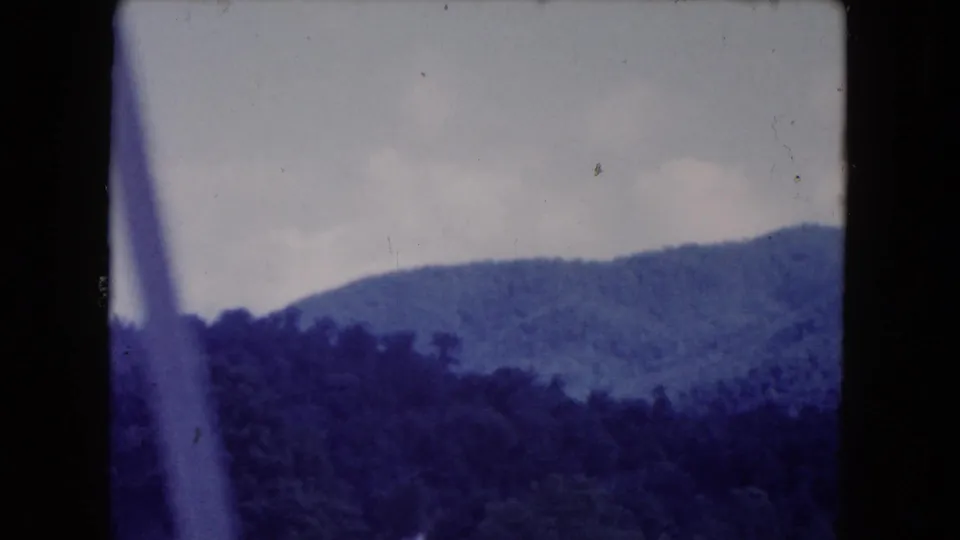 1960s 8mm travel footage shows forested hills passing behind a blurred vertical foreground bar, likely part of a window or vehicle frame.