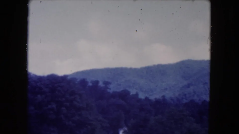 1960 8mm home-movie frame showing wooded hills under an overcast sky, filmed from a moving travel viewpoint.