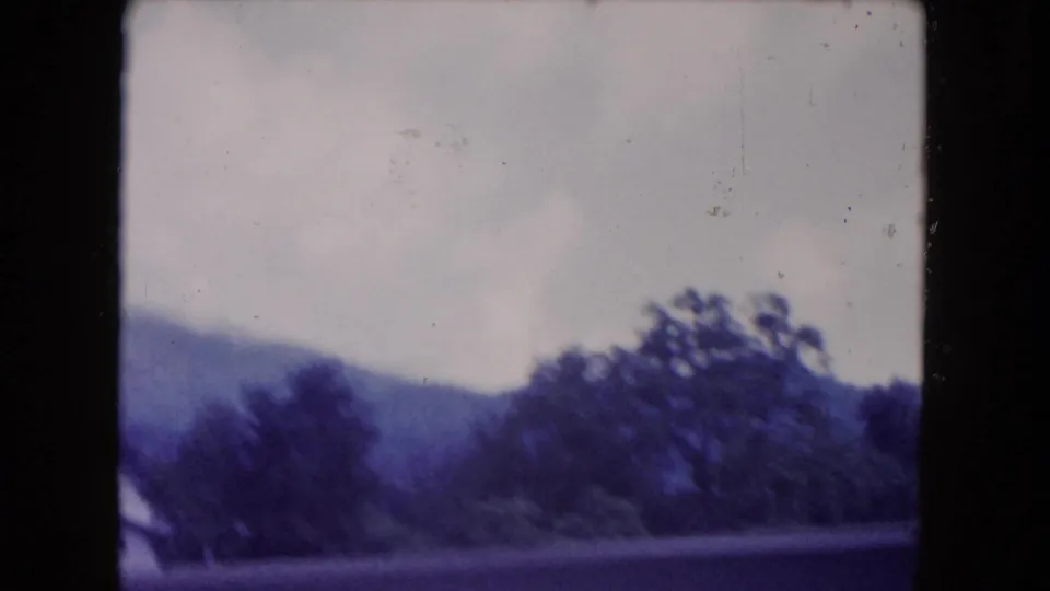 Soft 1960 8mm frame with distant hills and roadside trees viewed from a moving car, with heavy film borders visible.
