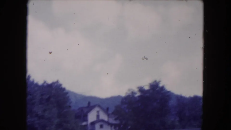 1960 8mm travel frame showing a light-colored house among trees with wooded hills in the background.
