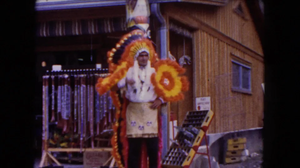 1960 8mm frame showing an adult in Native American-style costume outside a wooden roadside building with hanging merchandise.
