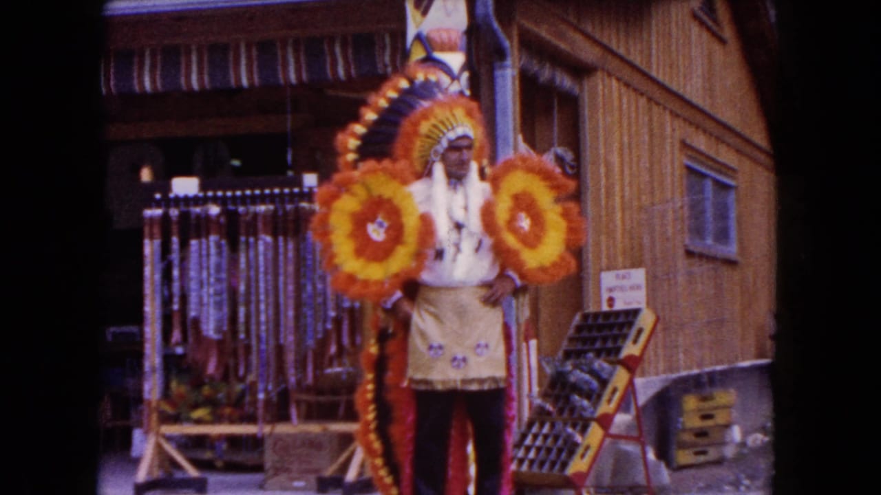 Costumed man in feathered headdress stands outside a wooden storefront with floral props.