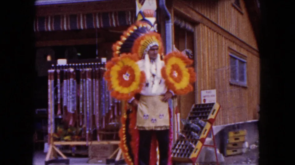 1960 Miami home-movie frame of a costumed man posing outside a wooden storefront with large orange and yellow floral props.