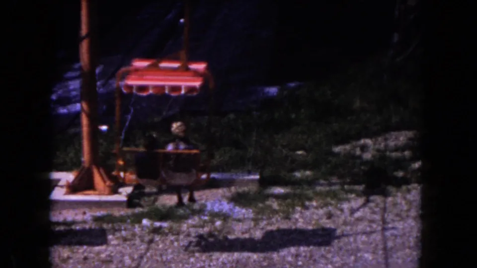 Nighttime 1960 Miami home-movie frame of a person seated on a red sky lift or chair ride over a dim outdoor attraction area.