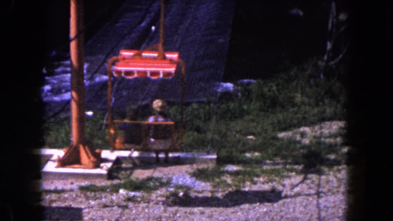 1960 Miami 8mm frame showing a red tram chair carrying two passengers past an orange support post and rocky green terrain.