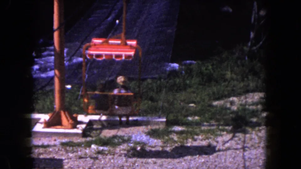 1960 Miami 8mm frame showing a red tram chair carrying two passengers past an orange support post and rocky green terrain.
