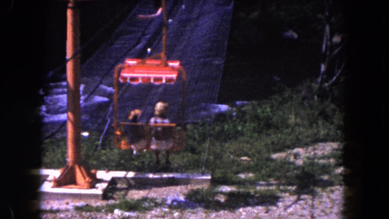 1960 Miami home-movie frame of two riders in a red tram chair passing above rocks, shrubs, and a shaded hillside.