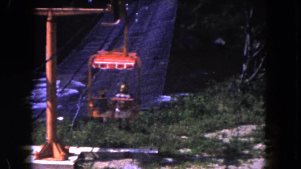 1960 Miami 8mm still with a red tram chair near an orange pole, framed by dark trees, brush, and rocky terrain.