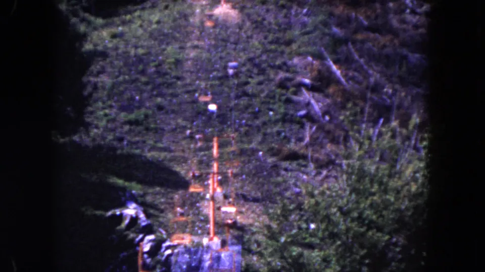 1960 8mm frame showing a wooded hillside with an orange ski-lift tower, cable line, and cleared ground.