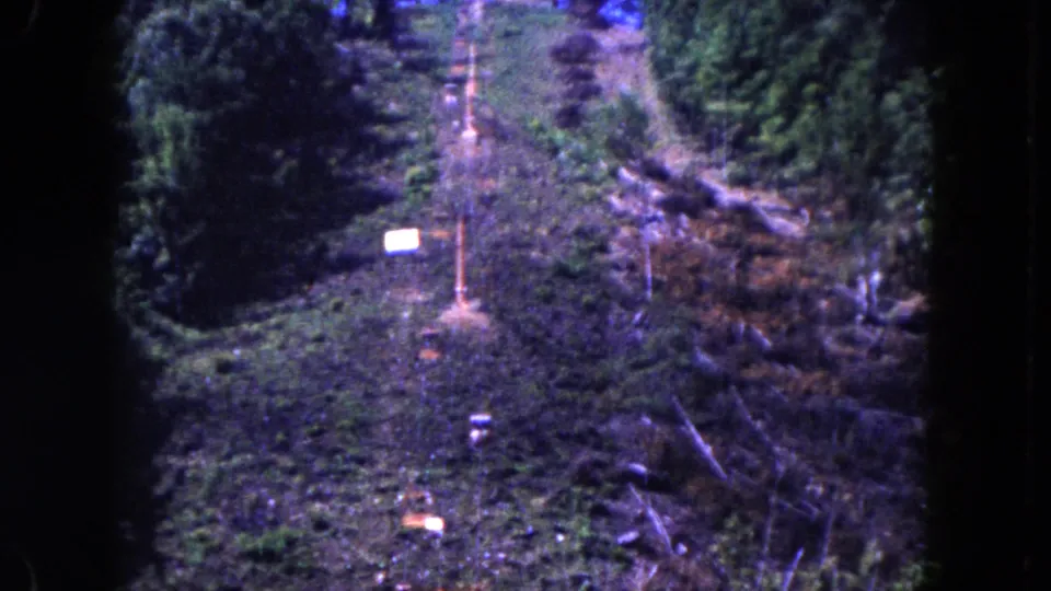 1960 8mm frame looking along a lift corridor on a wooded slope, with orange poles and cables visible.