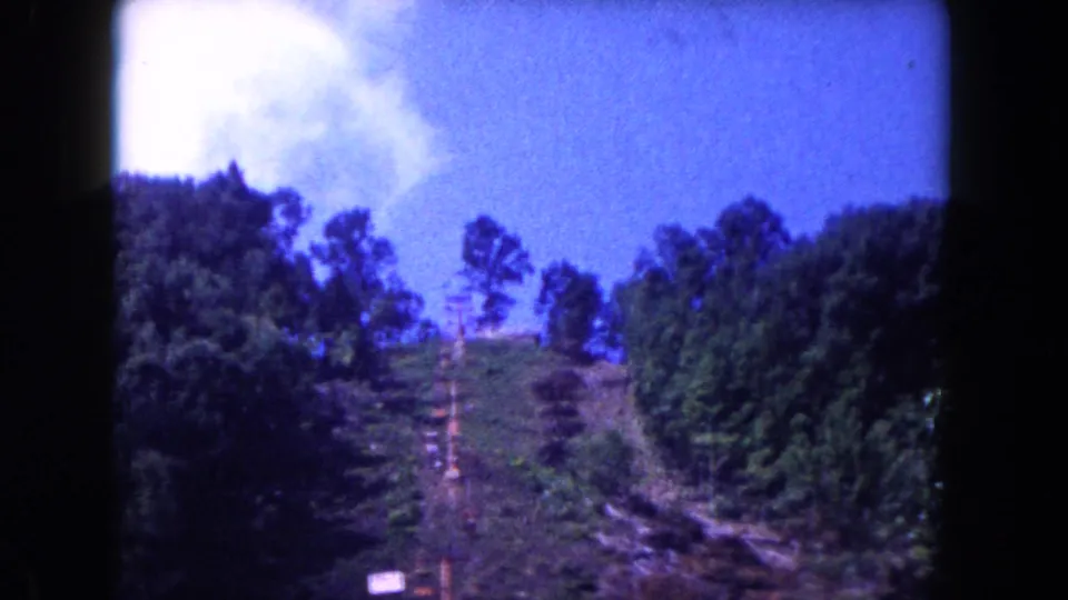 1960 Miami frame with the camera tilted slightly upward toward a wooded slope, blue sky, dirt path, and small pale sign.