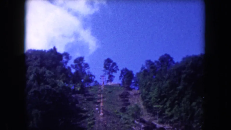 1960 8mm frame of a wooded hillside and blue sky, with soft focus and dark film borders.