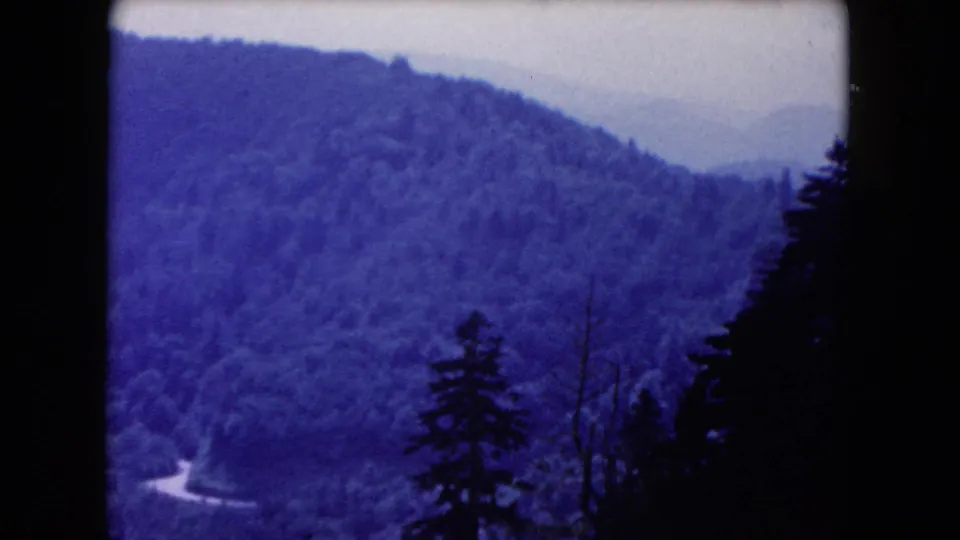 1960 8mm frame of blue-tinted wooded hills, distant ridges, a winding road segment, and foreground pine silhouettes.