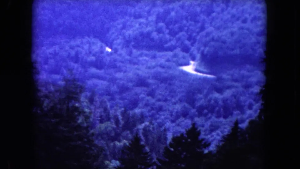 1960 8mm frame showing a blue-tinted wooded slope, pale winding road sections, and dark conifers in the foreground.