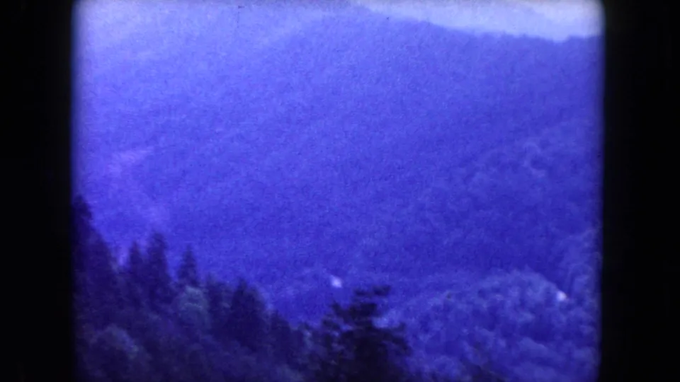 Soft 1960 8mm landscape frame with blue mountain ridges, dark foreground treetops, and a small pale road trace below.