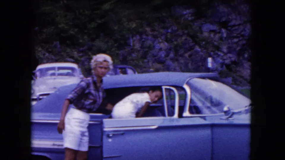 1960 home-movie frame of travelers beside a blue mid-century car at a roadside stop, with other parked cars and trees behind them.