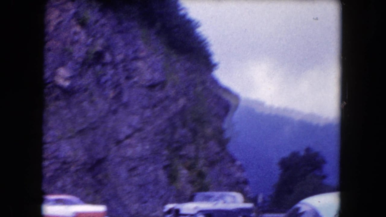 Soft 8mm frame showing automobiles along a curving road beside rocky cliffs, with mountains and cloudy sky in the distance.