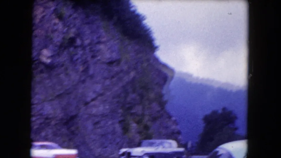 Soft 8mm frame showing automobiles along a curving road beside rocky cliffs, with mountains and cloudy sky in the distance.