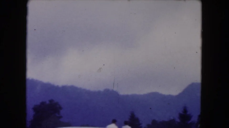 Landscape view from the home movie showing hazy mountains, cloudy sky, trees, and small roadside figures near the bottom of the frame.